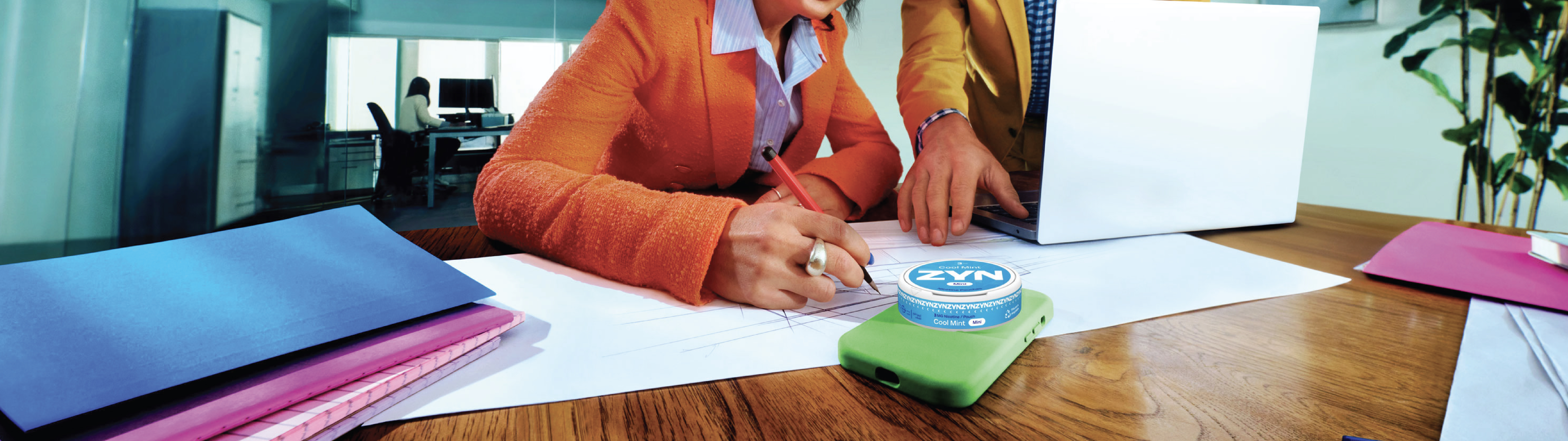 A woman in an orange blazer sitting at a desk writing on paper in front of a laptop with a man in a yellow blazer leaning over. A white ZYN nicotine pouch container is placed on top of a phone in a green case on the table.